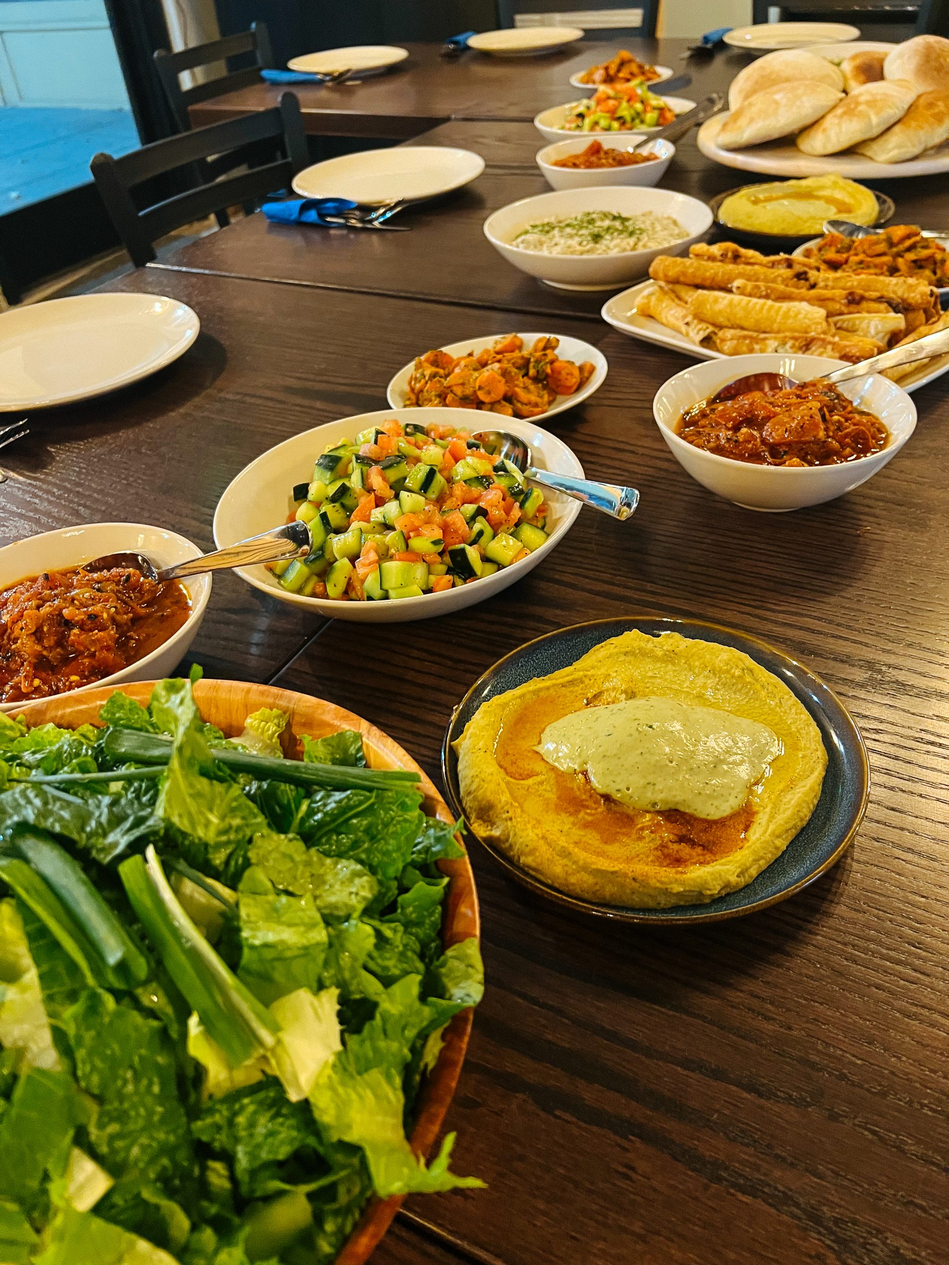 Dining table spread with bowls of chopped salad, dips, marinated vegetables, pastry rolls, and pita bread, leafy green backdrop, warm inviting tones