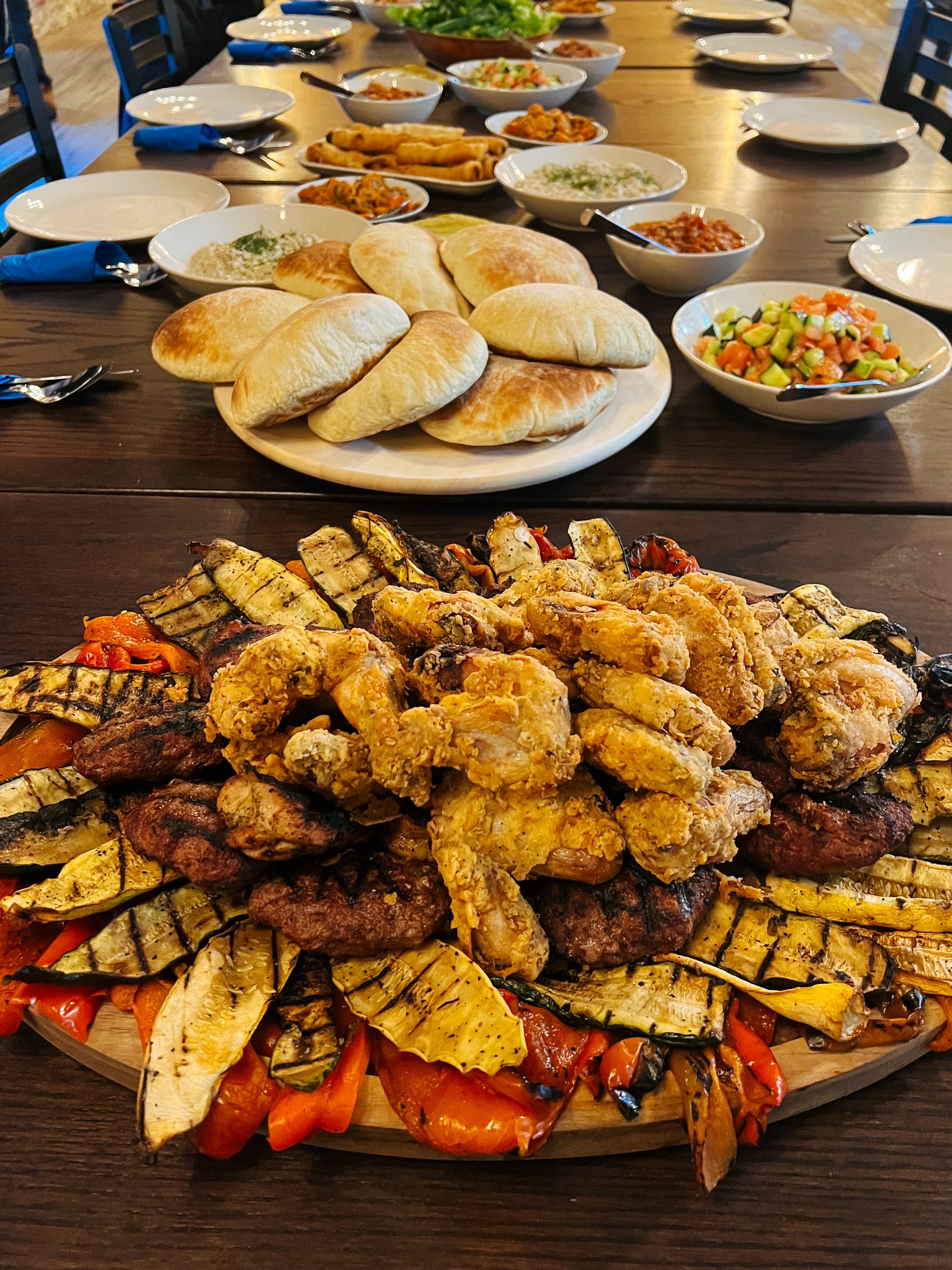 Wooden table with a large platter of grilled vegetables, fried chicken pieces, and meat patties in front, pita bread placed around, warm earthy tones