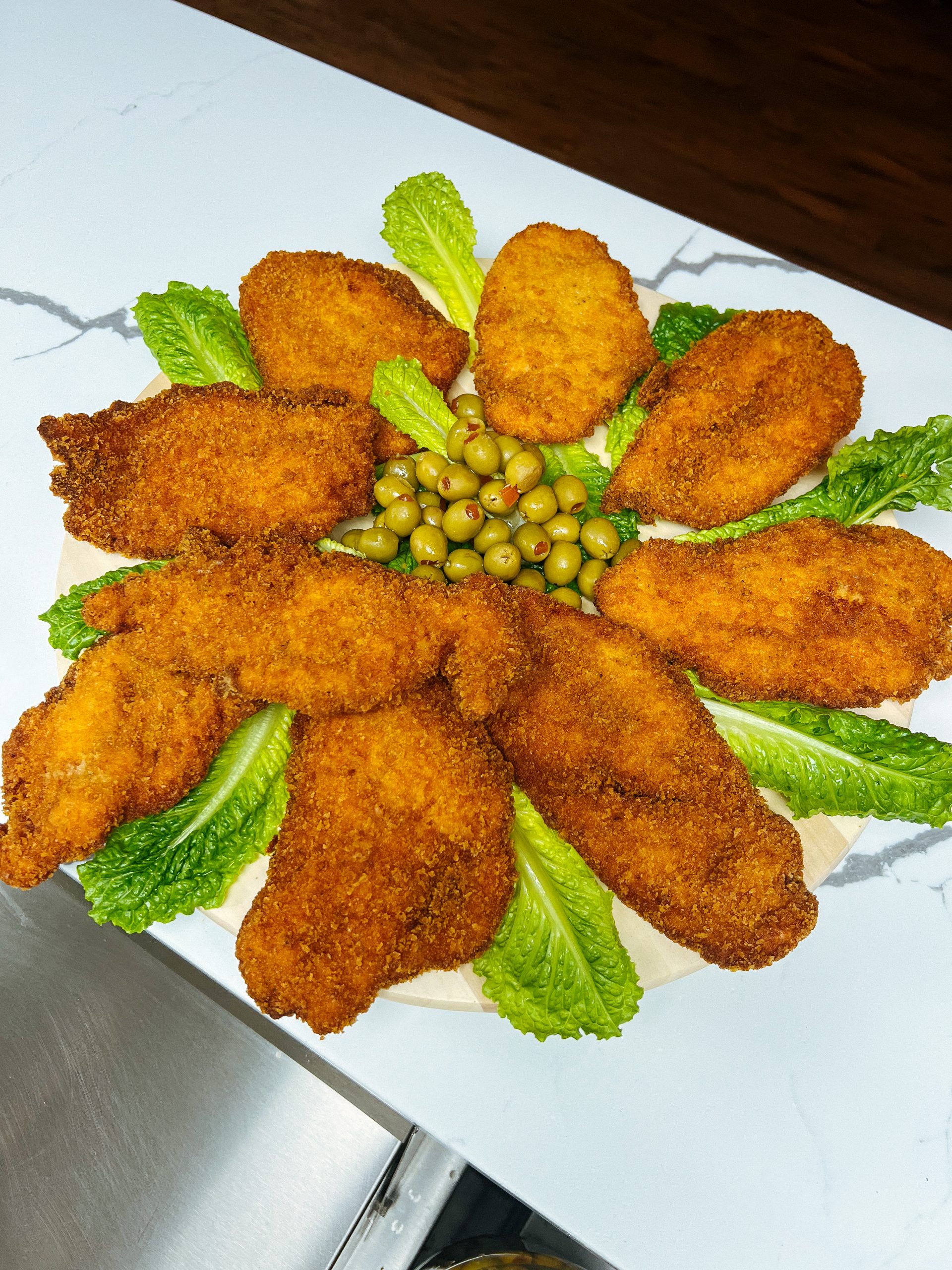Breaded fried cutlets arranged in a flower pattern on lettuce leaves with green olives in the center, on a marble counter