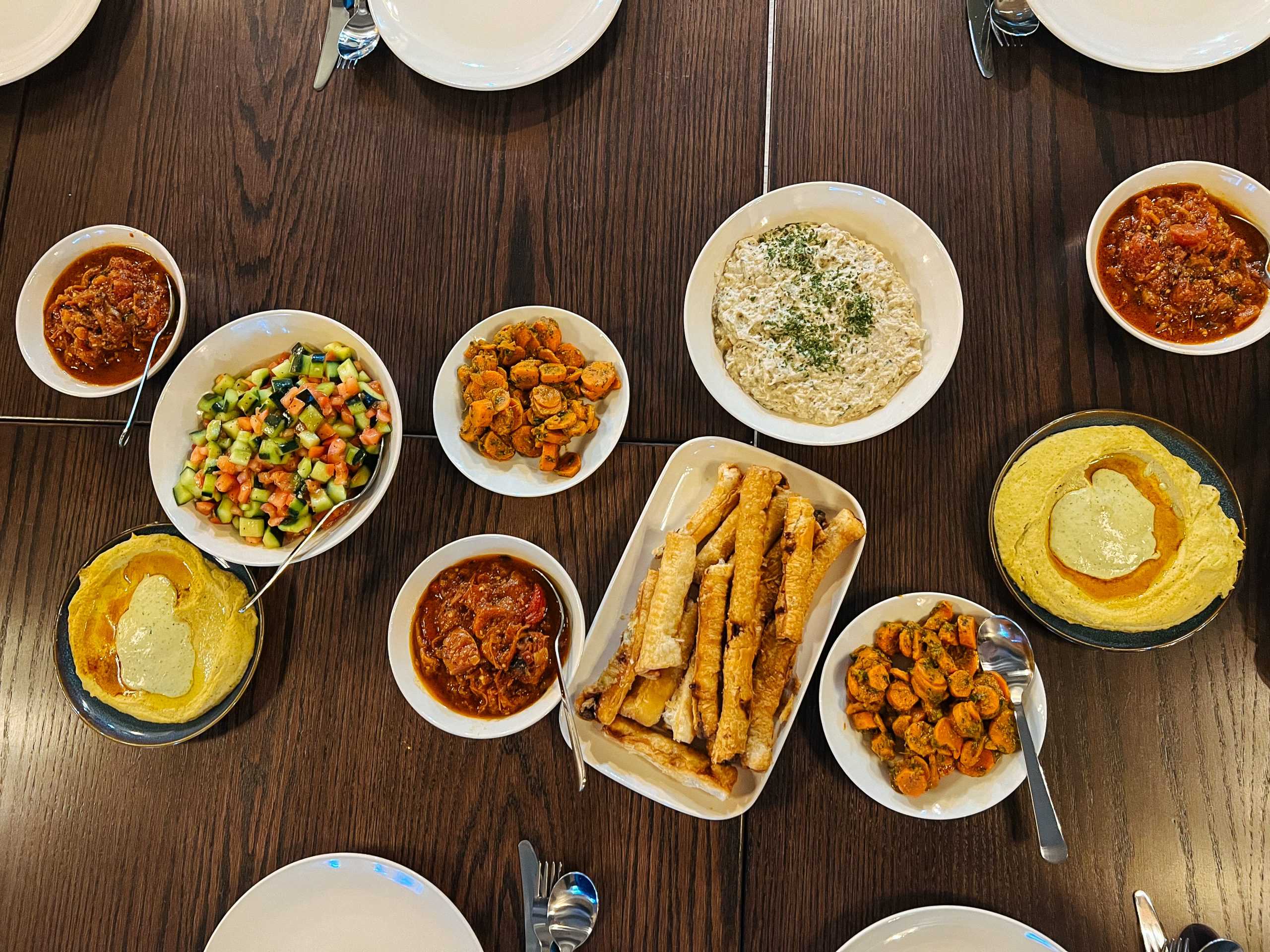 Overhead view of a wooden table filled with small bowls of dips, chopped salad, marinated carrots, pastry rolls, and hummus