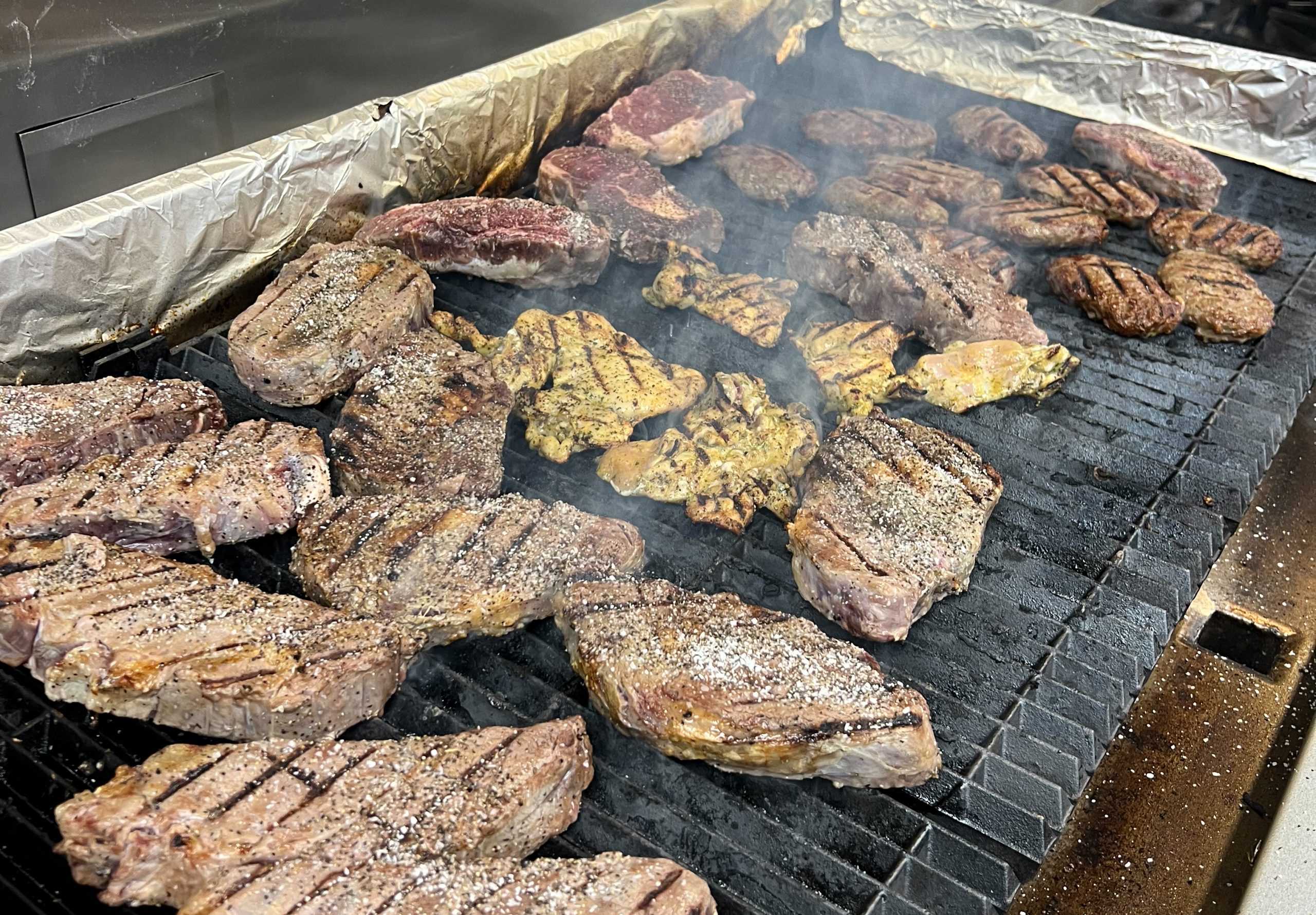 Assortment of steaks, chicken pieces, and meat patties grilling on a large outdoor grill with smoke and char marks, dark smoky tones